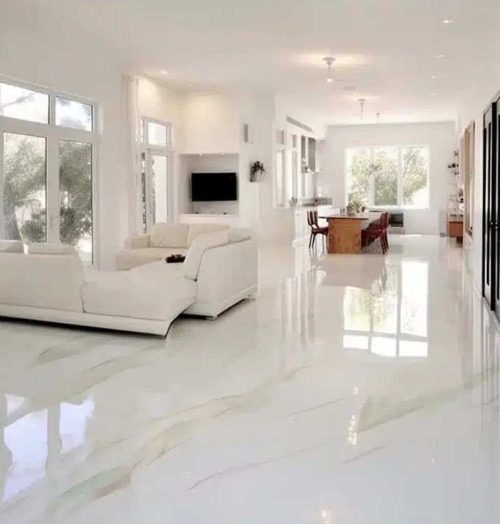 A bright, modern living space featuring a glossy white floor with subtle beige veining. The room has large windows allowing natural light, a white sectional sofa, a wooden dining table, and a flat-screen TV on the wall.